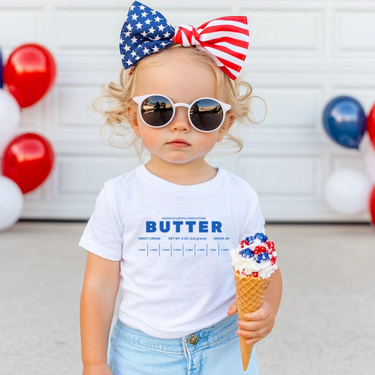 Toddler wearing white butter graphic tee styled with red, white, and blue accessories. Cute outfit idea for holidays, photos, and playful everyday wear.