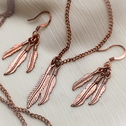 Copper tone feather jewelry set flat lay with dangle earrings and coordinating pendant necklace, western boho accessory set with detailed feather charms, nature inspired jewelry for women, lightweight earrings and statement necklace combination, gift for her perfect for festival outfits and everyday bohemian style