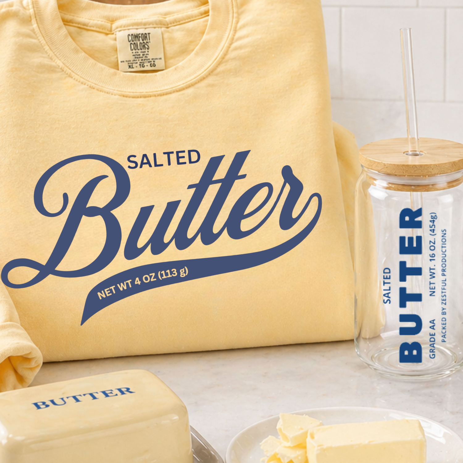 Close-up of butter-themed Zestful Productions products including a yellow “Salted Butter” sweatshirt, glass sipper jar, butter dish, and fresh butter styled on a modern kitchen countertop.