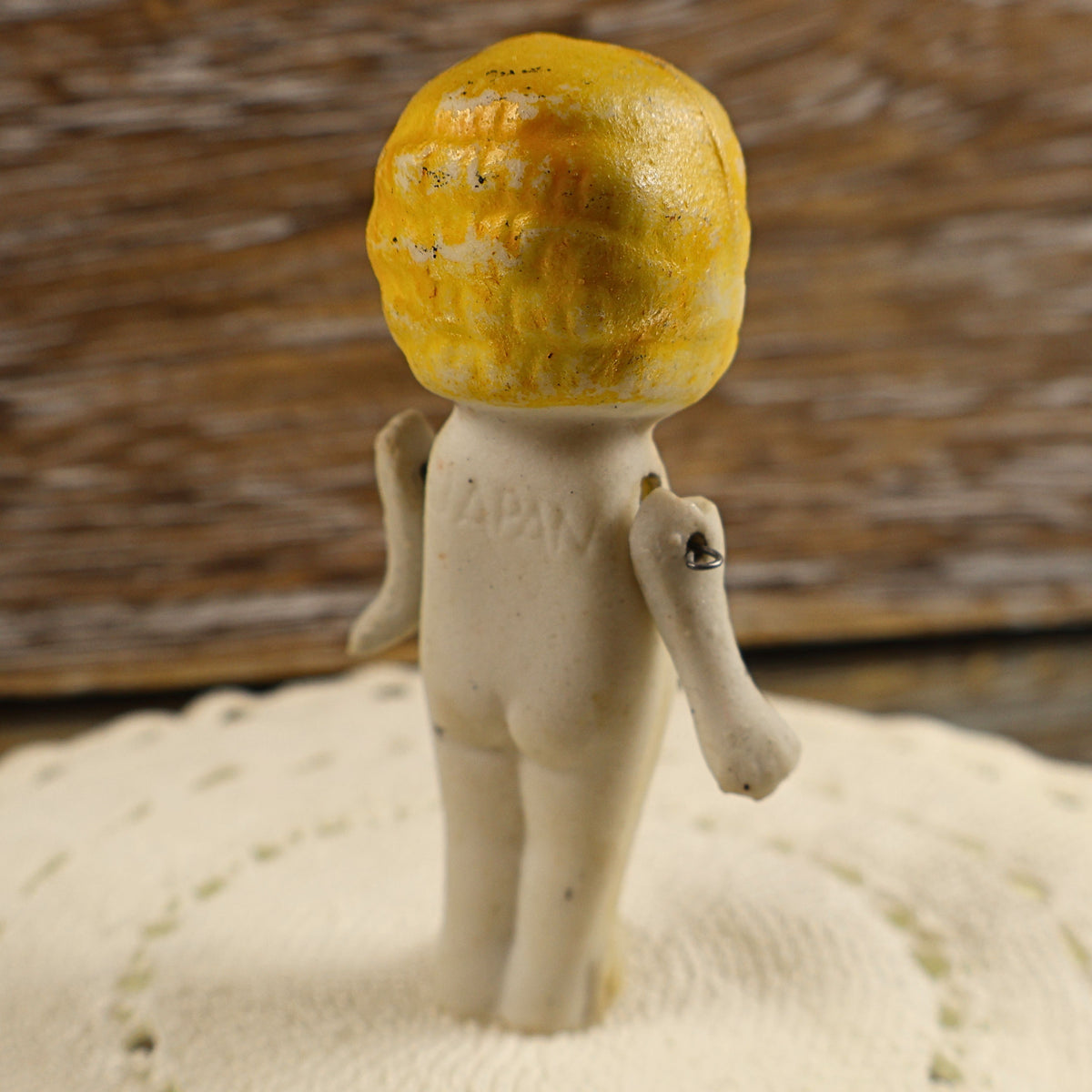 Rear view of the miniature bisque penny doll showing textured molded yellow hair, the matte ivory porcelain body, and an impressed JAPAN origin mark across the upper back. Small metal or wire fasteners connecting the movable arms are visible at both shoulders.