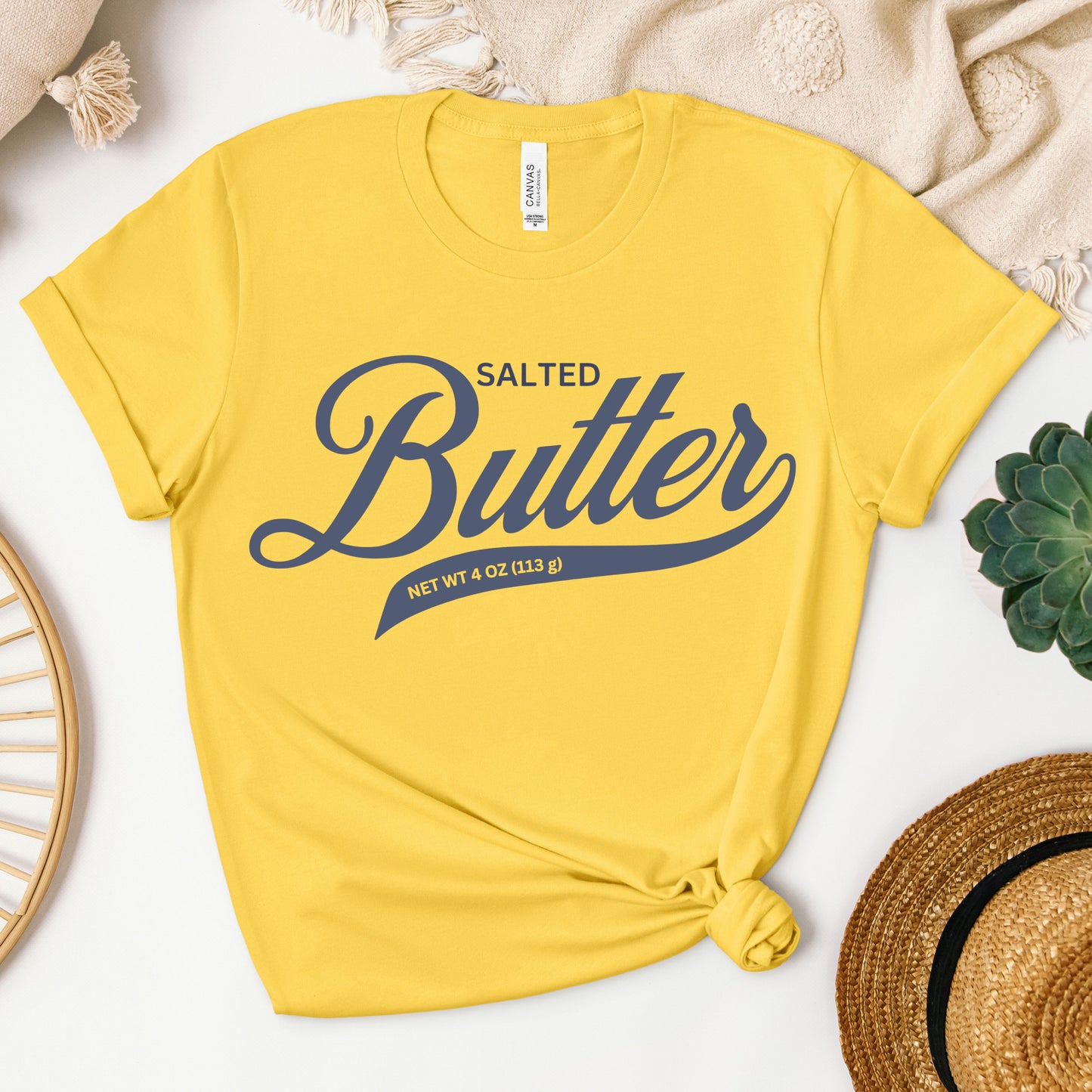 Yellow t-shirt with 'Salted Butter' text on a white surface with decorative items.