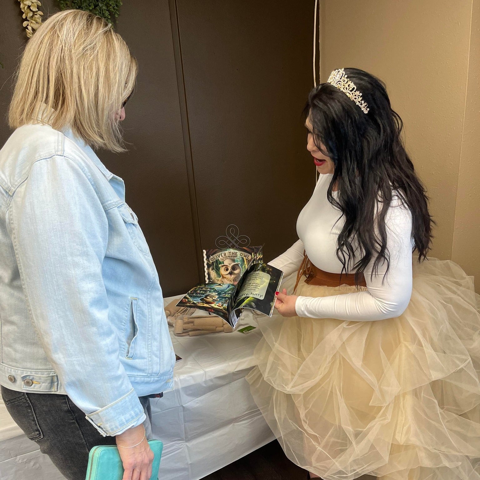 Woman in a white dress with a tiara interacting with another woman in a denim jacket.