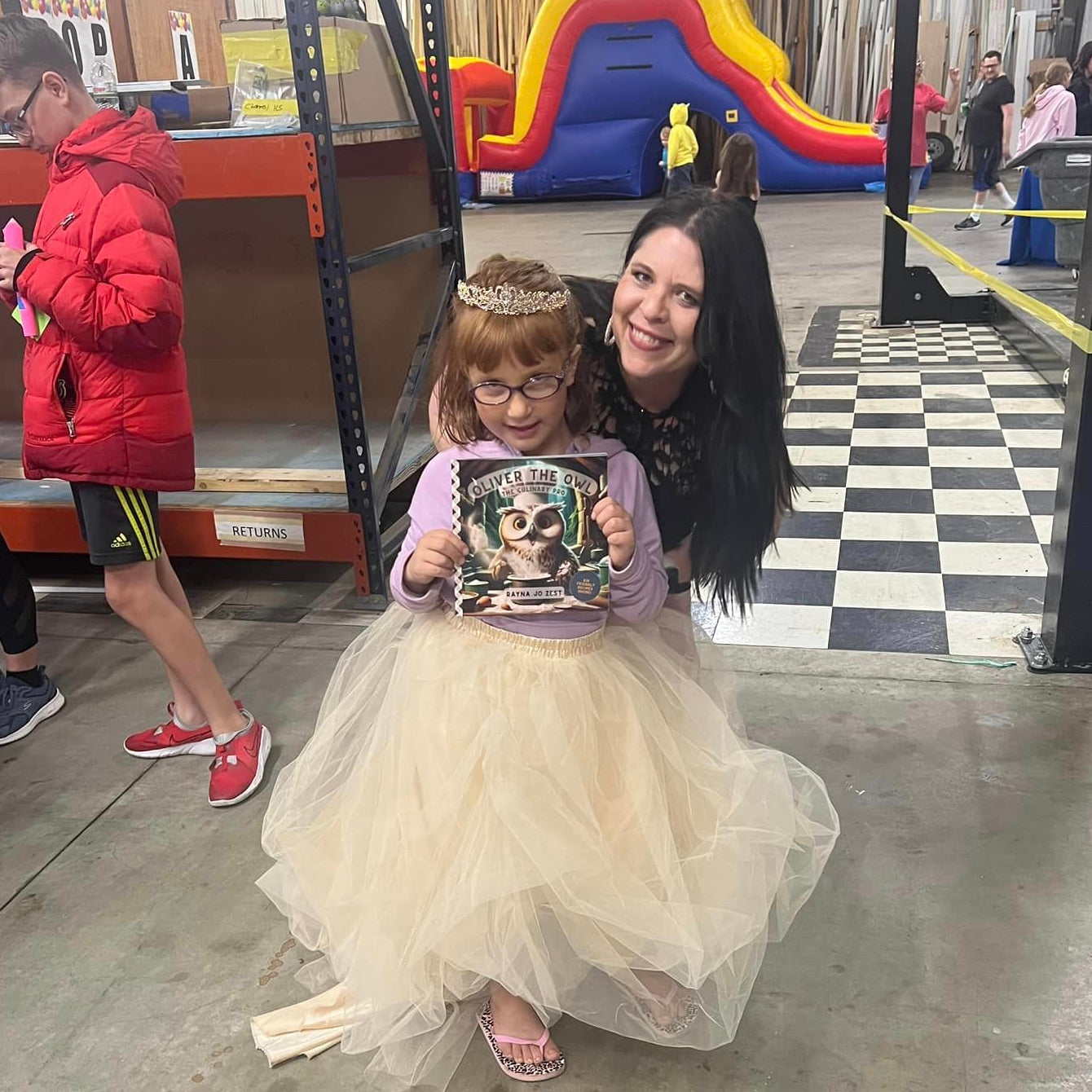 Woman and child in a costume at an indoor event with a checkered floor and inflatable slide in the background.