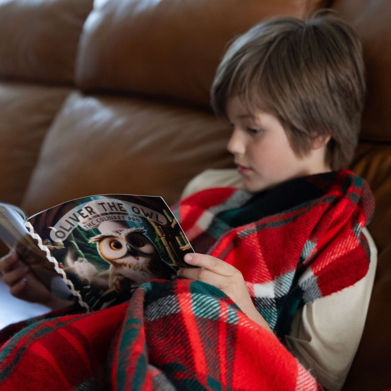 Child reading a book titled 'Oliver the Owl' on a brown couch with a red and green plaid blanket.