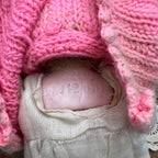 Close-up of the recessed markings on the back of the doll’s head, with the pink knitted clothing lifted and the cream garment visible below.
