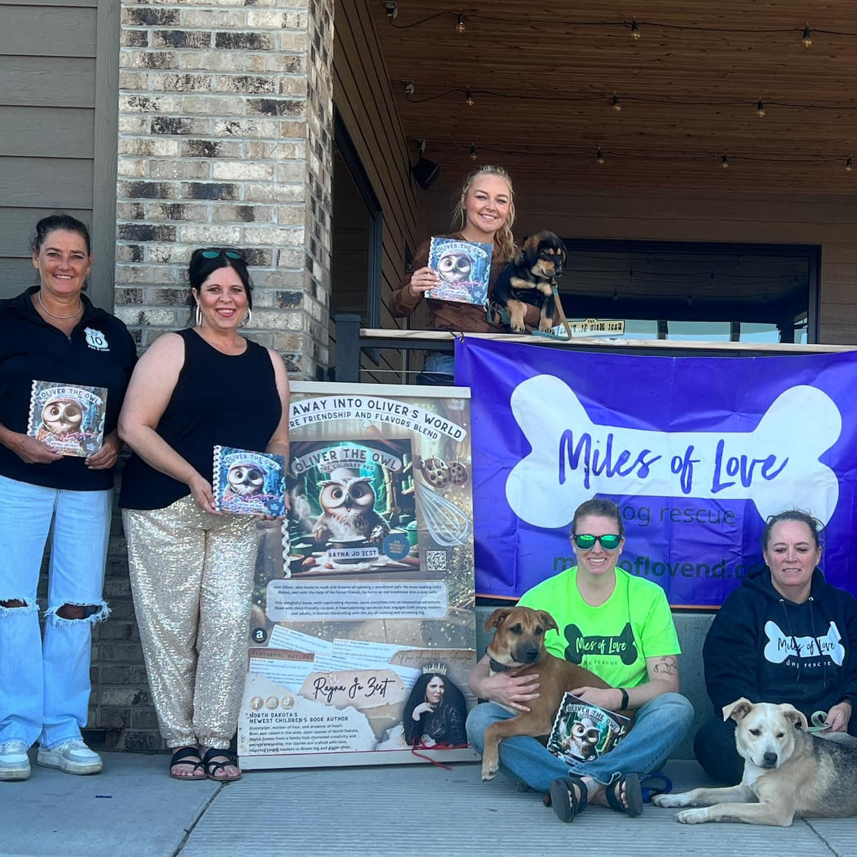 Group of people with dogs holding 'Oliver the Owl' books at a Miles of Love event outdoors.