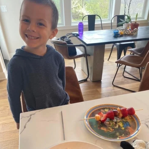 Child sitting at a table with a plate of skewered fruit in a home setting