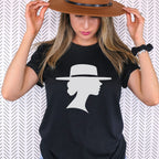 Woman holding the brim of a brown hat while wearing a black graphic tee featuring a light-colored silhouette of a woman in profile beneath a wide-brimmed hat.