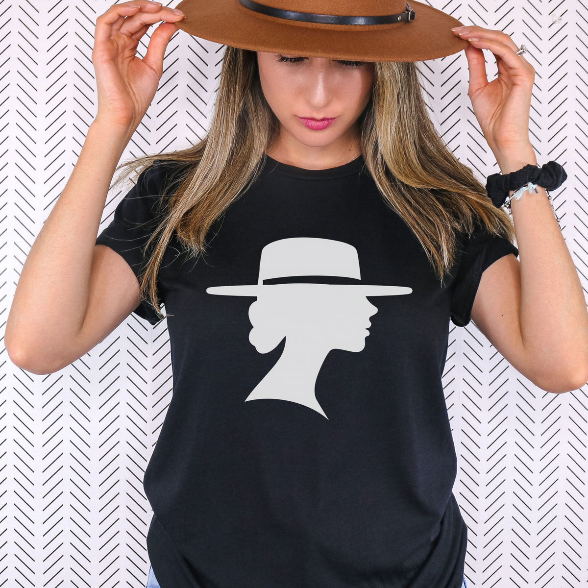Woman holding the brim of a brown hat while wearing a black graphic tee featuring a light-colored silhouette of a woman in profile beneath a wide-brimmed hat.