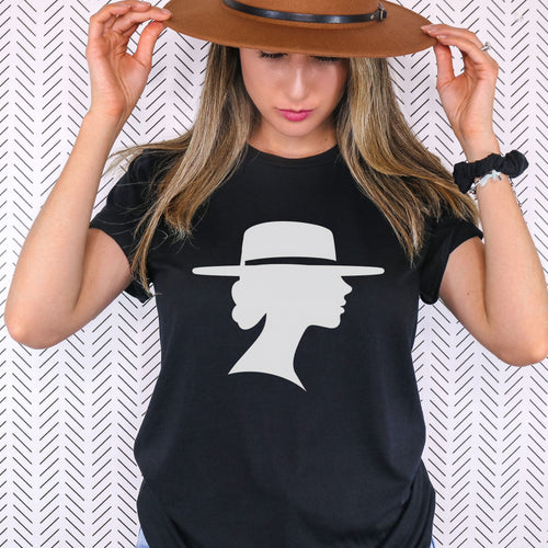 Woman holding the brim of a brown hat while wearing a black graphic tee featuring a light-colored silhouette of a woman in profile beneath a wide-brimmed hat.