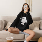 Woman seated on a beige sofa wearing a black sweatshirt with a light-colored graphic of a woman beneath a wide-brimmed hat, holding a mug near her face.
