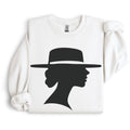 White crewneck sweatshirt arranged flat with folded sleeves, featuring a black silhouette of a woman in side profile beneath a wide-brimmed hat.