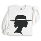 White crewneck sweatshirt arranged flat with folded sleeves, featuring a black silhouette of a woman in side profile beneath a wide-brimmed hat.