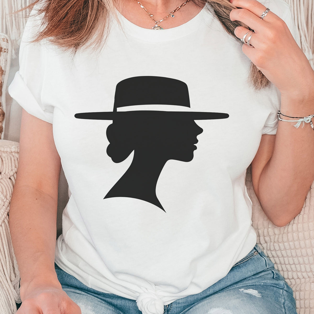 Close-up of a woman wearing a white graphic tee knotted at the waist with blue jeans. The design features a black silhouette of a woman in profile wearing a wide-brimmed hat.