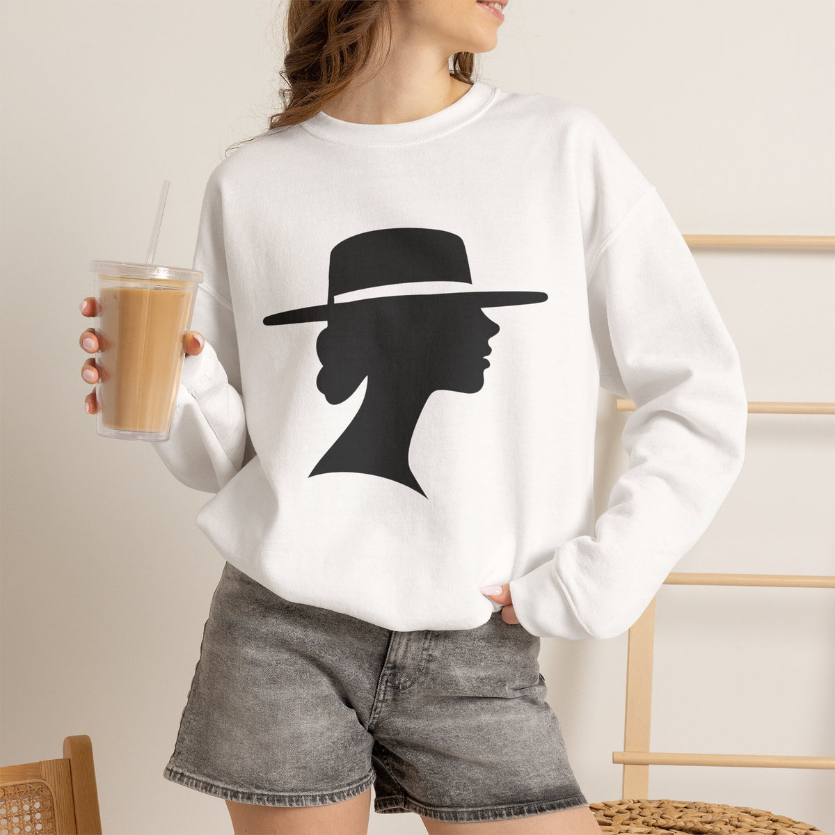 Woman holding a tall iced drink while wearing a white silhouette graphic sweatshirt with gray denim shorts, with one hand tucked into a pocket.