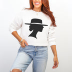 Woman wearing a white crewneck sweatshirt with light blue distressed jeans and gold jewelry. A black graphic depicts a woman in profile wearing a wide-brimmed hat.