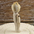 Side and back condition view of an antique Japanese penny doll with an unglazed ivory bisque body. The photograph shows the molded hairstyle, faded red bow, surface discoloration, darkened shoulder wire, rough residue near one arm joint, and age-related wear.