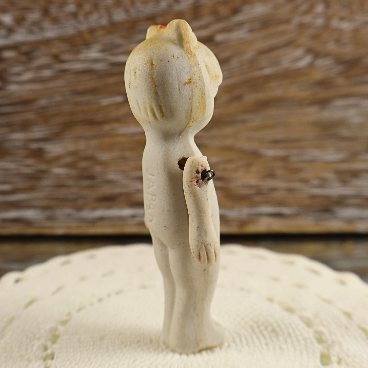 Side and back condition view of an antique Japanese penny doll with an unglazed ivory bisque body. The photograph shows the molded hairstyle, faded red bow, surface discoloration, darkened shoulder wire, rough residue near one arm joint, and age-related wear.