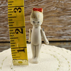 Vintage Japanese bisque penny doll standing beside a yellow measuring tape indicating a height of approximately three inches. The front-facing miniature has a molded red hair bow, painted eyes and lips, slim ivory bisque body, fixed legs, and wire-attached arms.