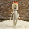 Front view of an antique Japanese bisque penny doll standing on cream crocheted fabric against a rustic wood background. The miniature girl has a matte ivory body, painted dark eyes, a molded hairstyle with a worn red bow, and separate arms attached at the shoulders.
