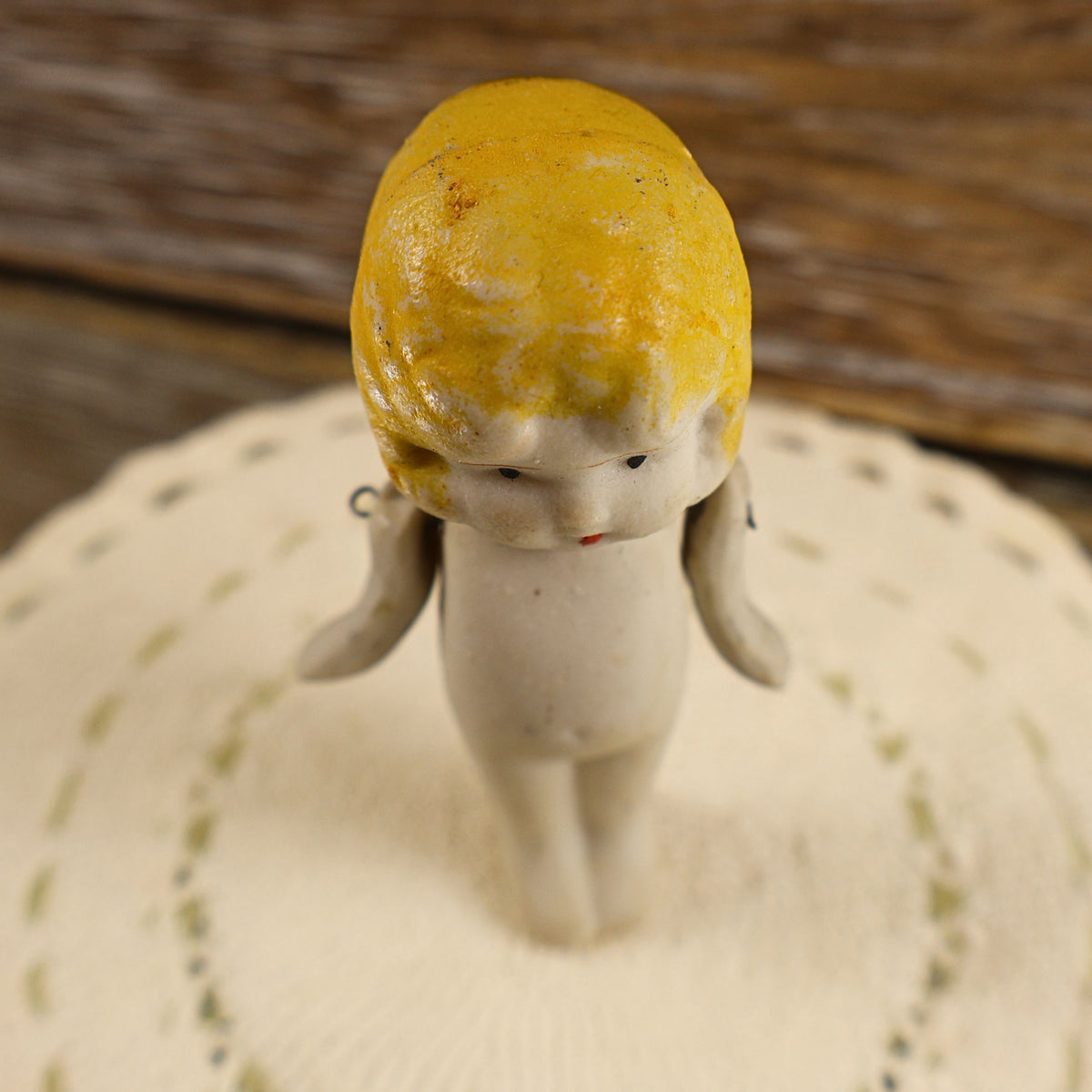 Overhead view of a small Japanese bisque doll with her head tilted slightly downward. The photograph shows the worn golden-yellow paint across the molded hairstyle, finely painted eyebrows and eyes, petite attached arms, and narrow stationary legs.