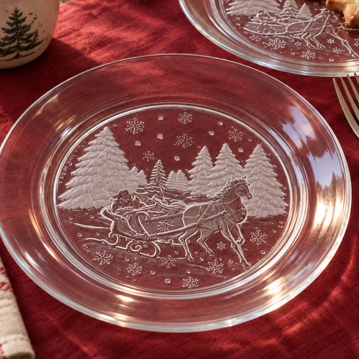 Illustrative holiday table mockup featuring clear glass sleigh-scene plates on cranberry fabric. An empty plate fills the foreground, with a second plate holding cookies behind it, alongside a tree-patterned mug and a partially visible silver fork.