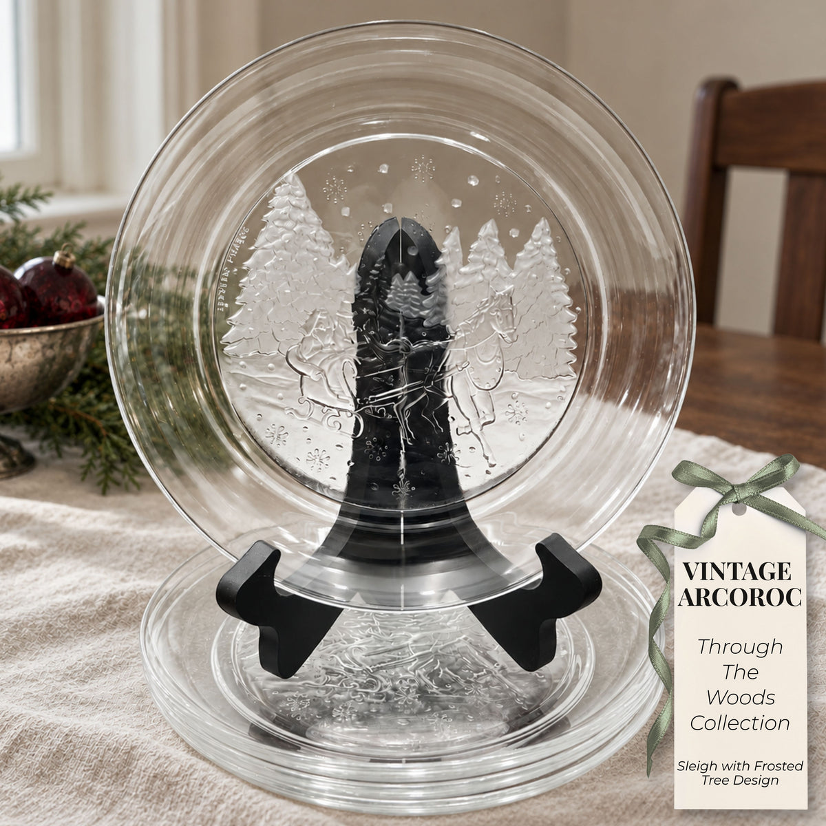 Styled holiday mockup showing an Arcoroc Through the Woods plate upright on a black stand above stacked glass plates. Ivory linen, greenery, burgundy ornaments, and a decorative product tag surround the display beside a window and wooden chair.