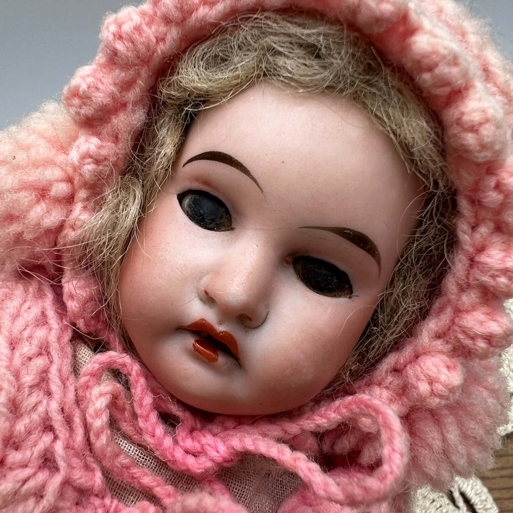 Close-up of the antique doll’s face framed by a pink knit bonnet, showing dark inset eyes, painted eyelashes and eyebrows, pink cheeks, parted lips, and light-colored hair.