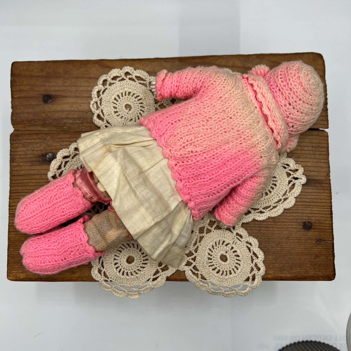Full-length rear view of the doll lying on a lace doily, showing the back of her pink knit bonnet and cape, cream skirt, and pink knitted footwear.