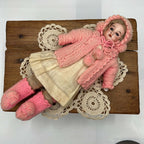 Full-length front view of the antique doll lying on a lace doily, wearing a pink knit bonnet and cape over a cream dress, with pink knitted footwear.
