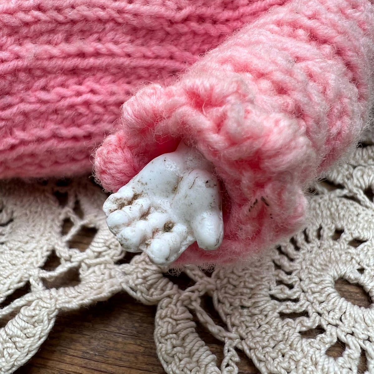 Detail of one pale doll hand extending from a pink knitted sleeve, showing the molded fingers and visible surface condition against a lace doily.