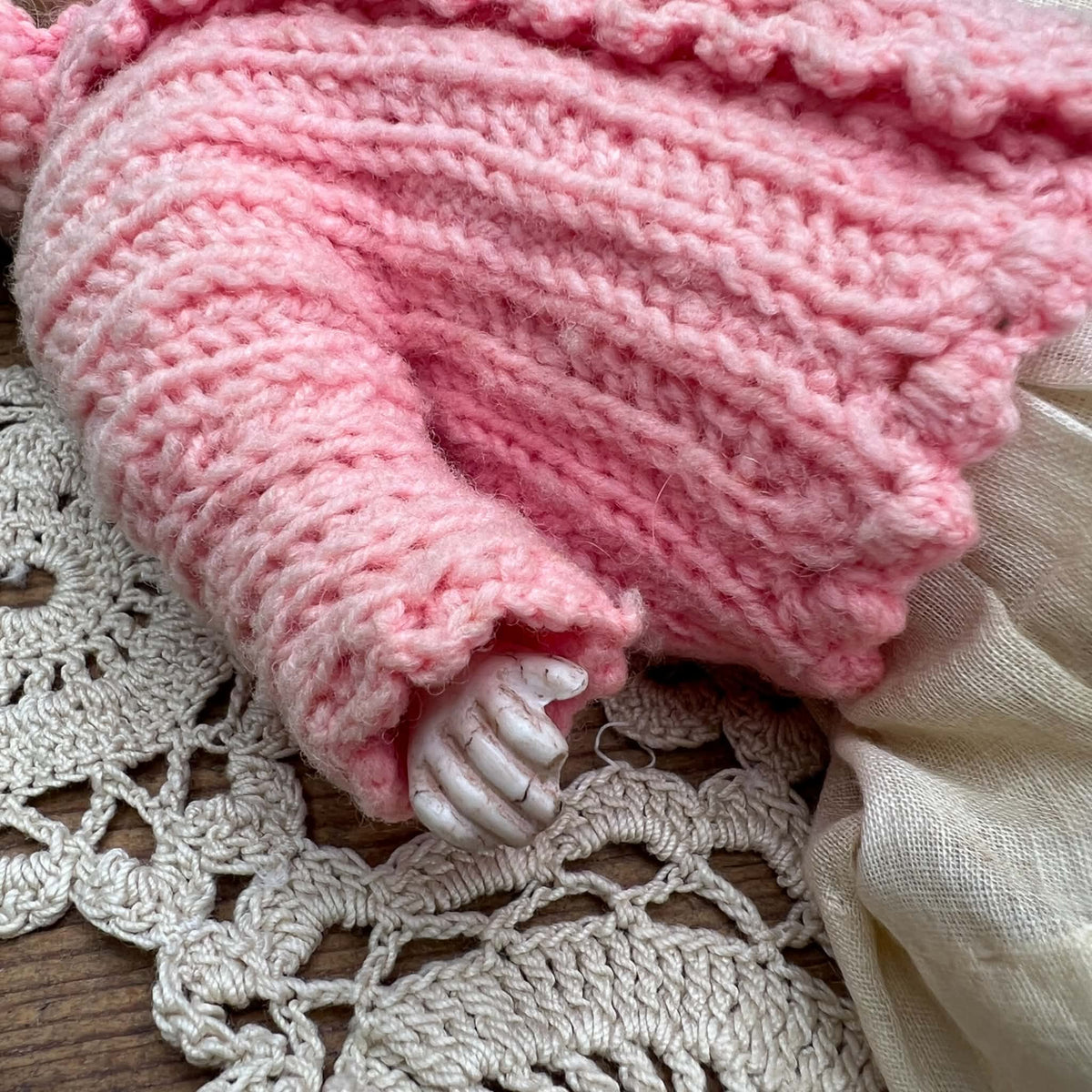 Close-up of the doll’s other hand beneath a pink knitted cuff, showing the fingers, surface coloring, and surrounding knit texture.