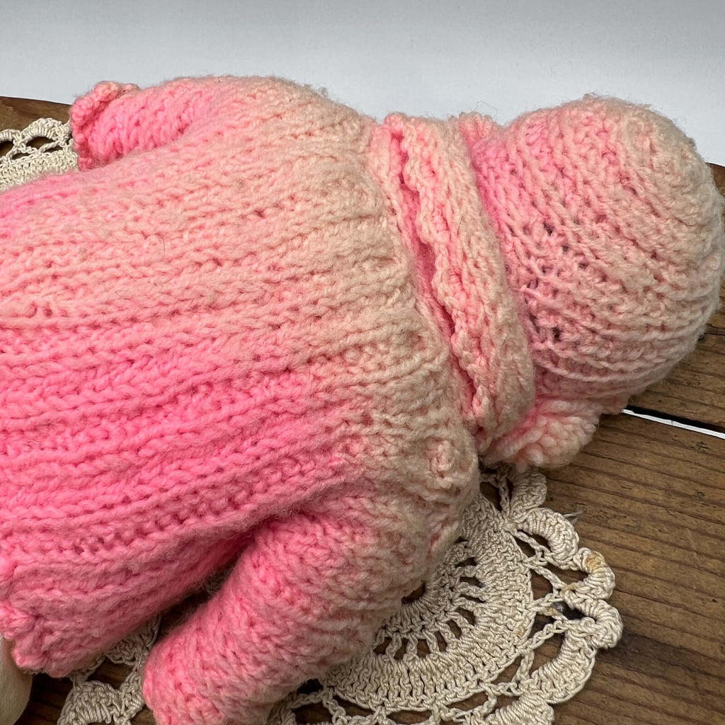 Close-up of the back of the doll’s pink knitted cape and bonnet, showing the stitch patterns, gathered shaping, and contrasting shades of pink yarn.
