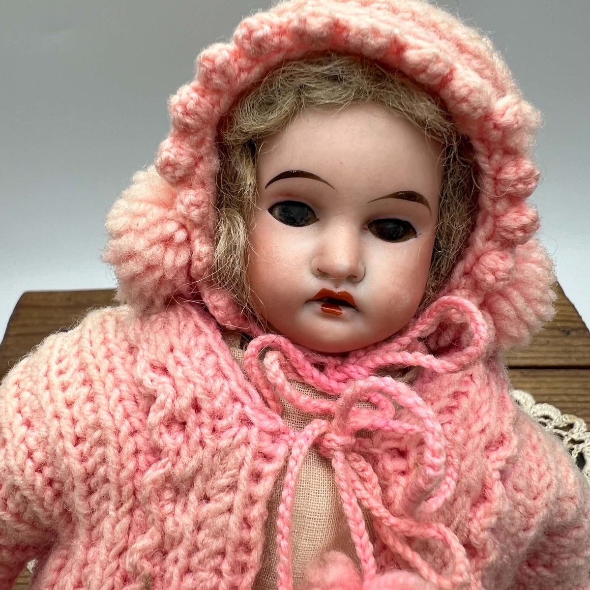 Front portrait of an antique Armand Marseille doll wearing a pink knit bonnet and matching cape tied beneath her chin, with dark eyes, painted brows, rosy cheeks, and an open mouth.