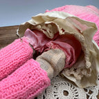 View beneath the doll’s lifted cream skirt, showing pink underclothing, fabric-covered legs, and one pink knitted foot covering beside an uncovered foot.