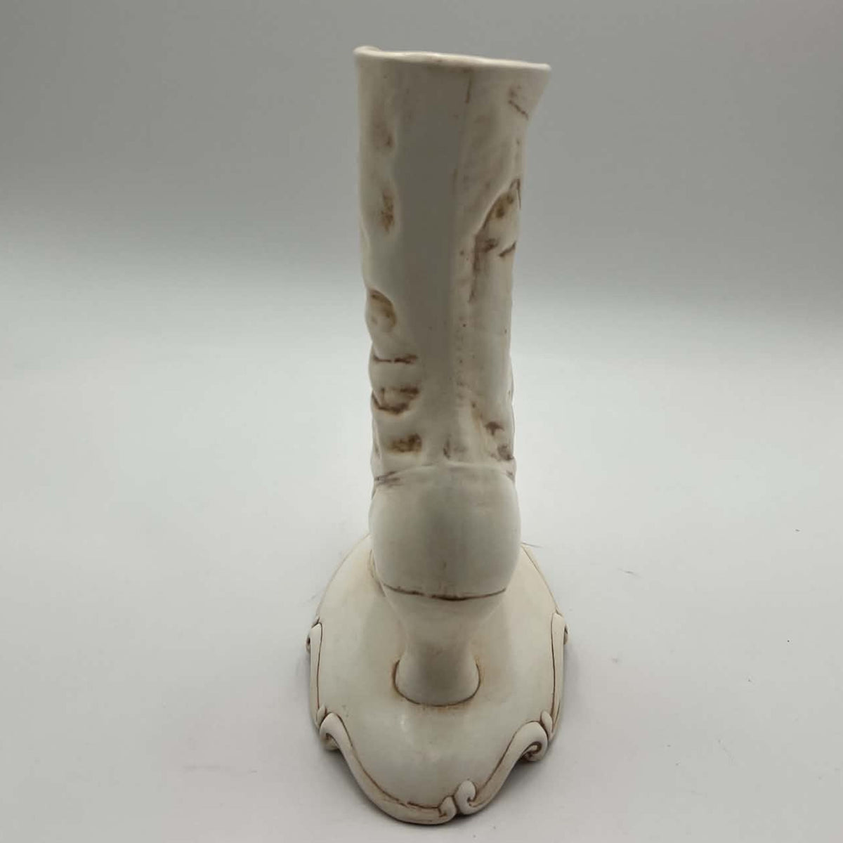 Rear view of a tall vintage Victorian boot vase showing the smooth back of the ceramic shaft, sculpted folds and small heel rising above an ornate pedestal base. Visible finish variations and brown distressed accents contribute to its hand-decorated vintage character.