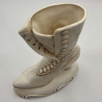 Three-quarter view of a vintage Atlantic Mold Victorian boot vase with a long toe, raised buttoned panel and ornate molded base. The substantial cream ceramic planter is photographed against a plain backdrop so its shape, finish variations and decorative details remain visible.