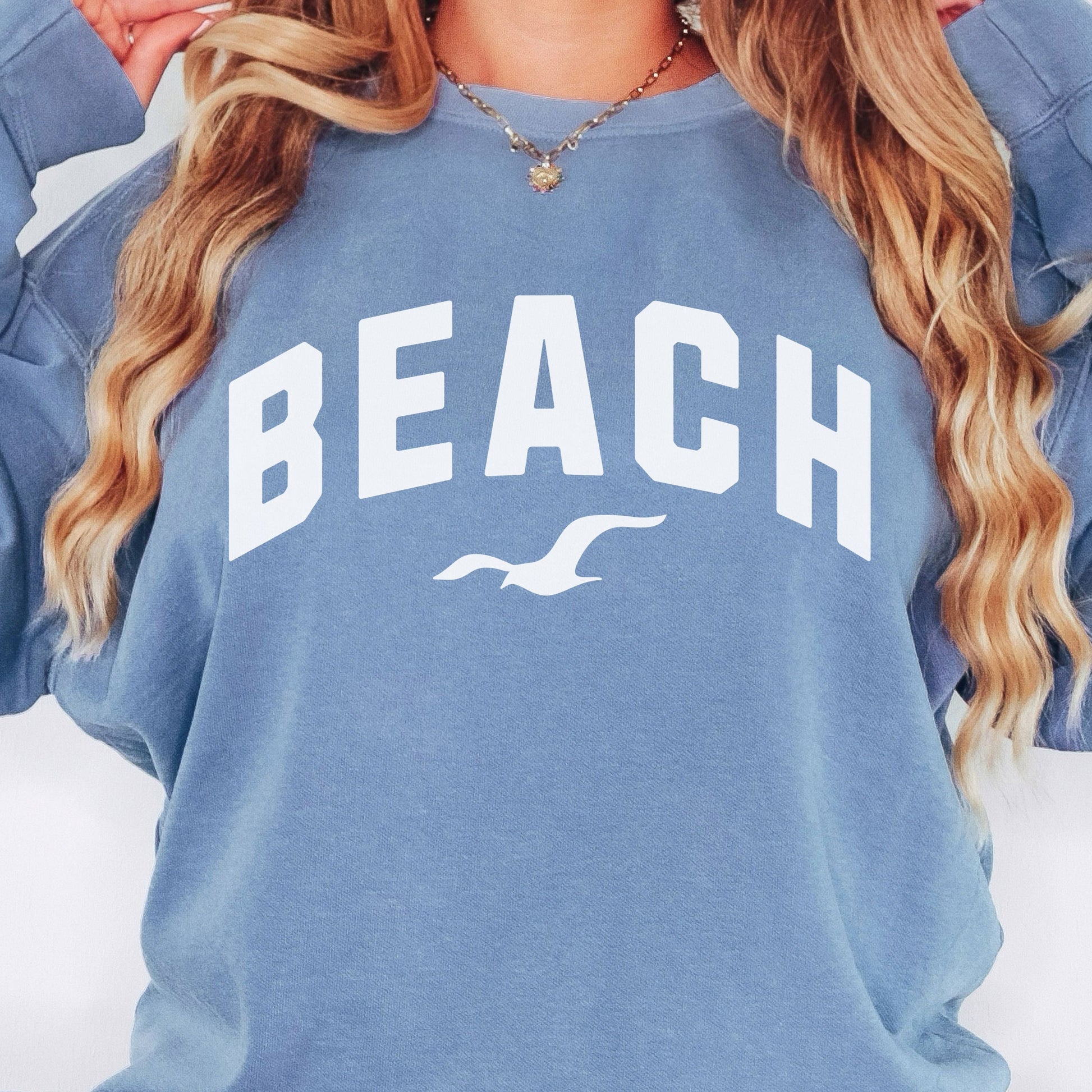 Blue jean beach crewneck sweatshirt close up on woman, oversized fleece pullover with beach graphic, soft fabric texture and relaxed coastal outfit detail