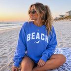 Flo blue beach crewneck sweatshirt worn by woman sitting cross legged on sand, relaxed oversized fleece pullover with beach graphic, cozy coastal vacation outfit
