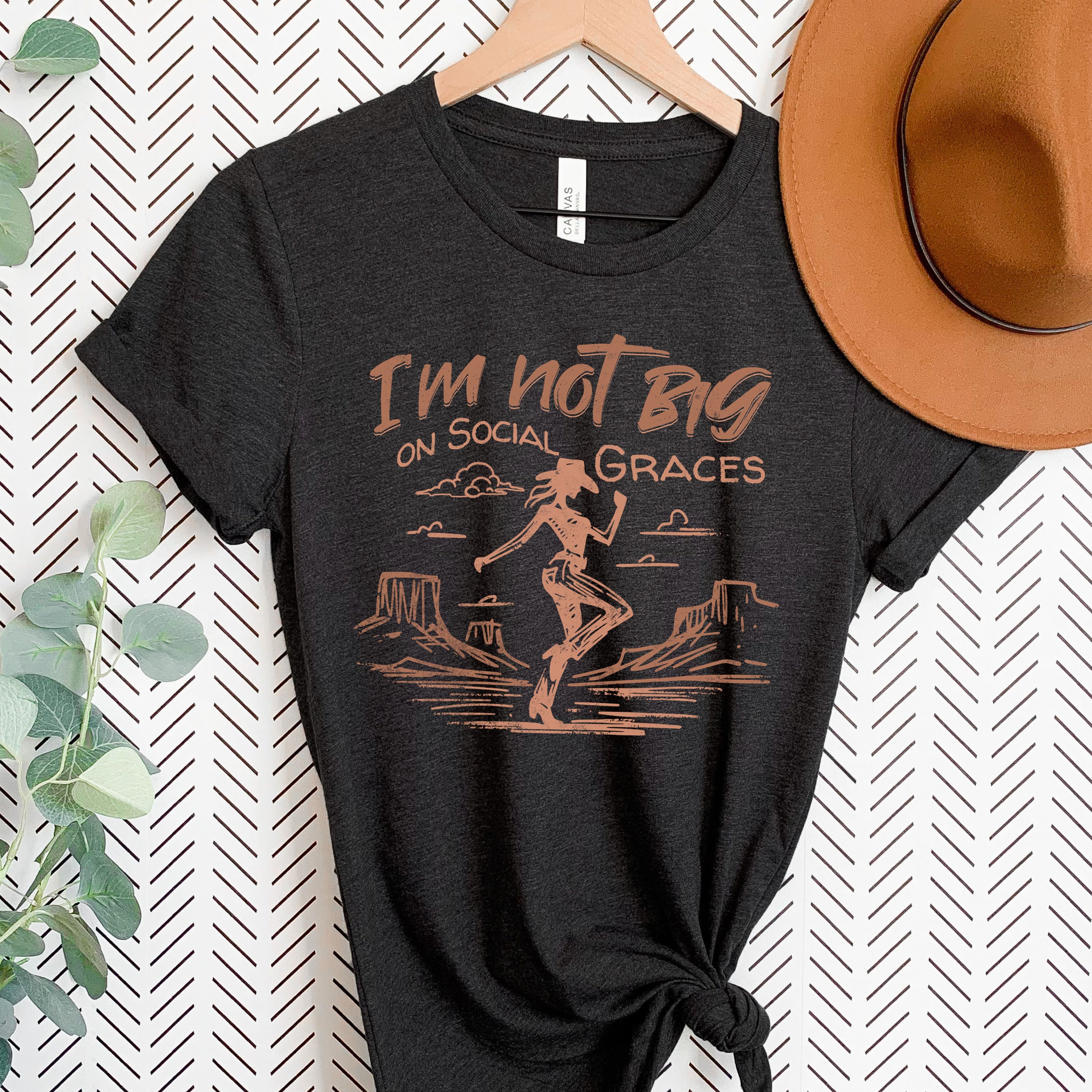 Black western graphic tee hanging with cowboy hat styling, featuring vintage cowgirl design and neutral copper print for a bold boutique look