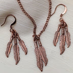 Feather jewelry set styled on neutral fabric featuring copper tone necklace and matching dangle earrings, western boho style jewelry with nature inspired feather pendant, lightweight statement necklace and earrings set, gift for her ideal for festival fashion, rustic outfits and layered bohemian accessories