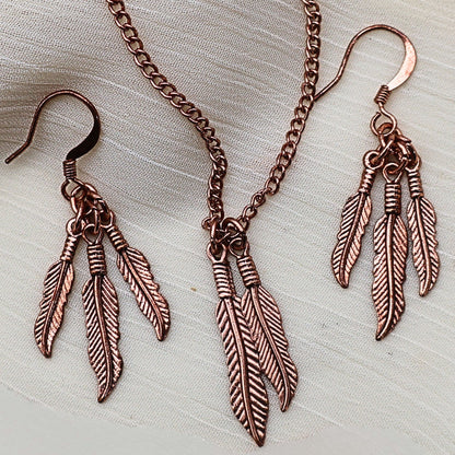Feather jewelry set styled on neutral fabric featuring copper tone necklace and matching dangle earrings, western boho style jewelry with nature inspired feather pendant, lightweight statement necklace and earrings set, gift for her ideal for festival fashion, rustic outfits and layered bohemian accessories