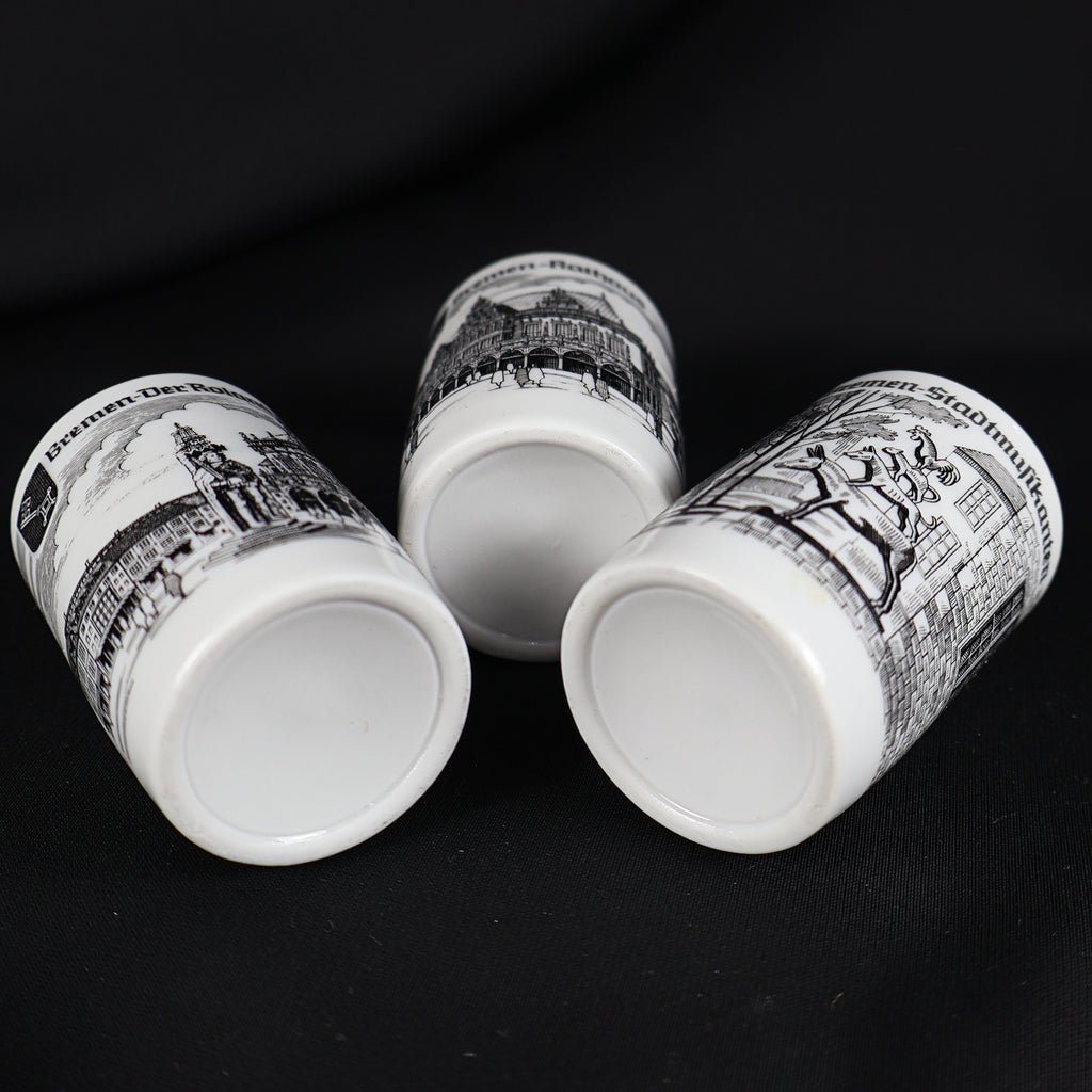Low angled view of three vintage Bremen Germany porcelain shot glasses resting on their sides against black fabric. Their unmarked recessed bases are prominently visible, along with portions of the Roland, Rathaus, and Bremen Town Musicians landmark illustrations.