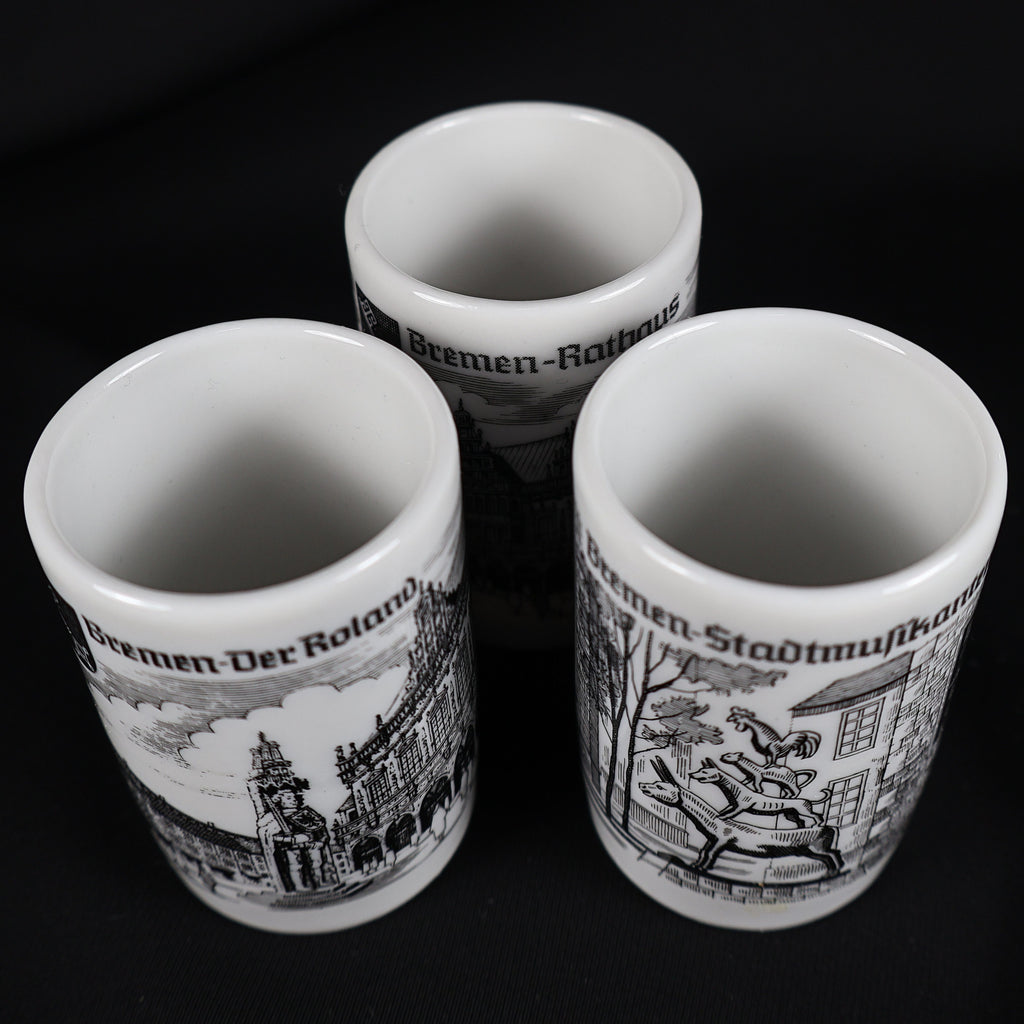 Overhead view into the three vintage white porcelain Bremen shot glasses, showing their clean cylindrical interiors and rounded rims. The exterior illustrations of the Roland statue, Town Musicians, and Rathaus remain partly visible against the solid black backdrop.