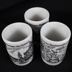 Overhead view into the three vintage white porcelain Bremen shot glasses, showing their clean cylindrical interiors and rounded rims. The exterior illustrations of the Roland statue, Town Musicians, and Rathaus remain partly visible against the solid black backdrop.