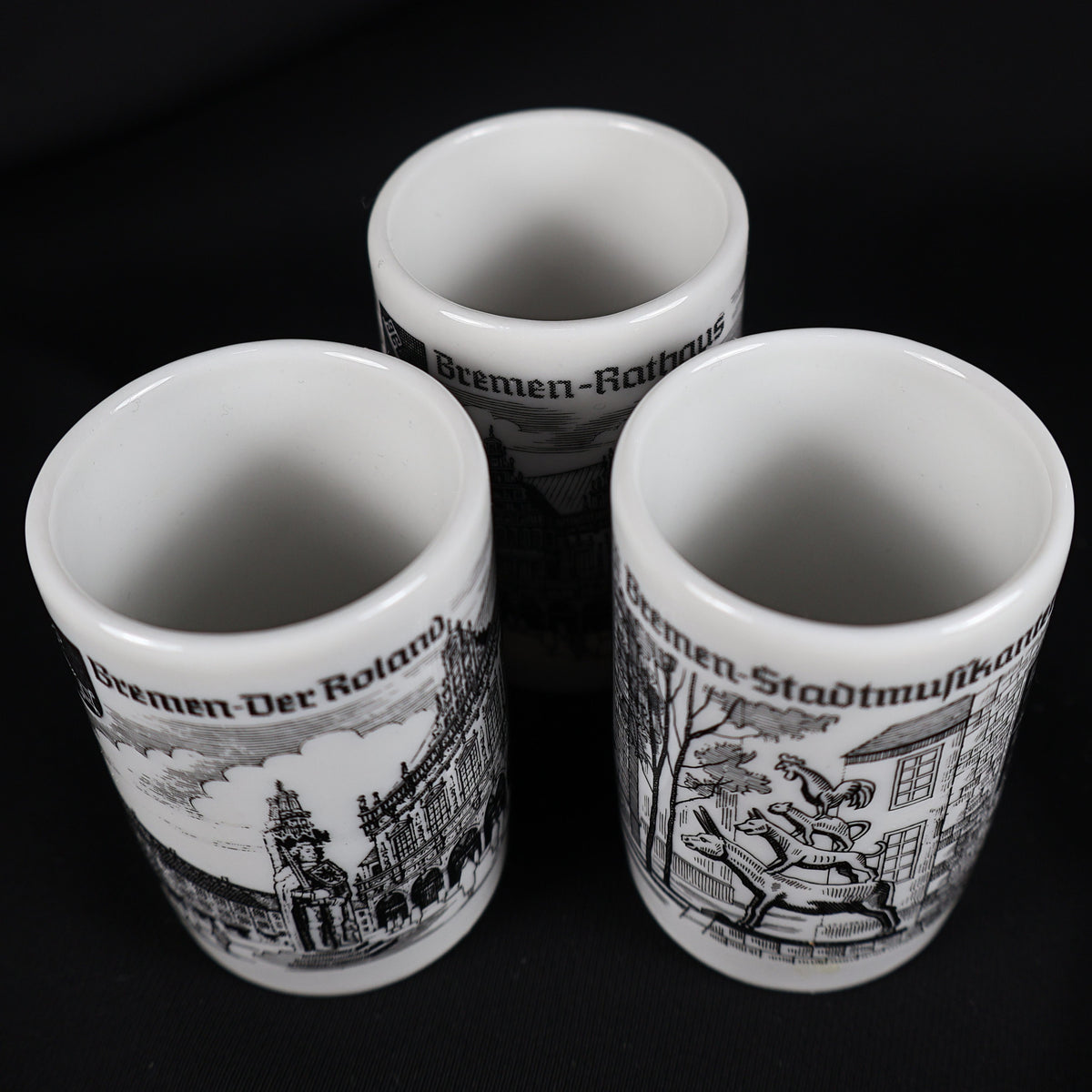 Overhead view into the three vintage white porcelain Bremen shot glasses, showing their clean cylindrical interiors and rounded rims. The exterior illustrations of the Roland statue, Town Musicians, and Rathaus remain partly visible against the solid black backdrop.