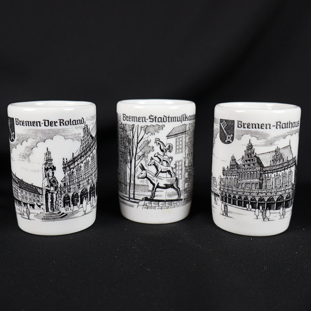 Front view of three coordinating white porcelain Bremen souvenir shot glasses displayed in a row. Black illustrations show the Roland statue beside historic buildings, the stacked Bremen Town Musicians, and the ornate Rathaus, with German titles above each scene.