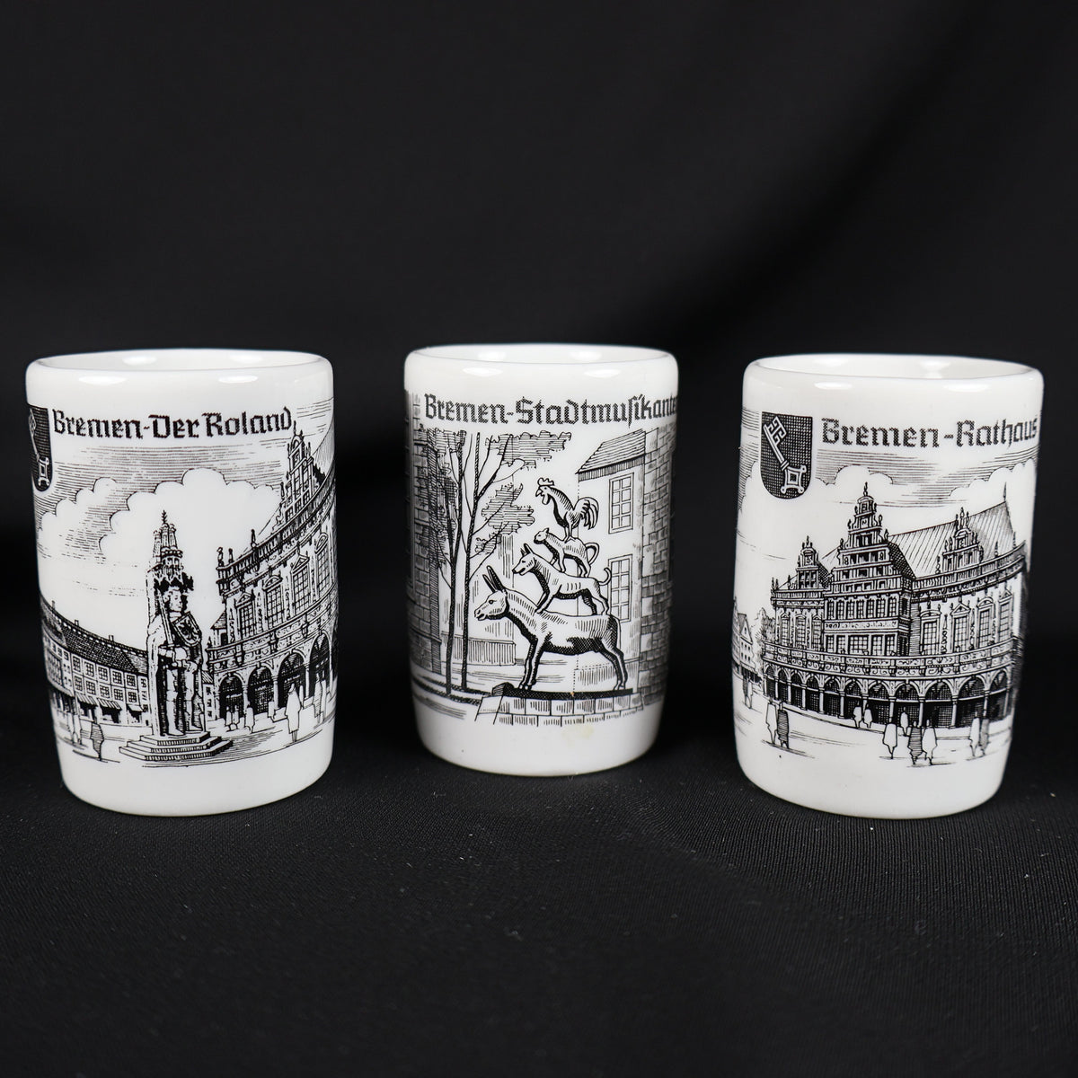 Front view of three coordinating white porcelain Bremen souvenir shot glasses displayed in a row. Black illustrations show the Roland statue beside historic buildings, the stacked Bremen Town Musicians, and the ornate Rathaus, with German titles above each scene.
