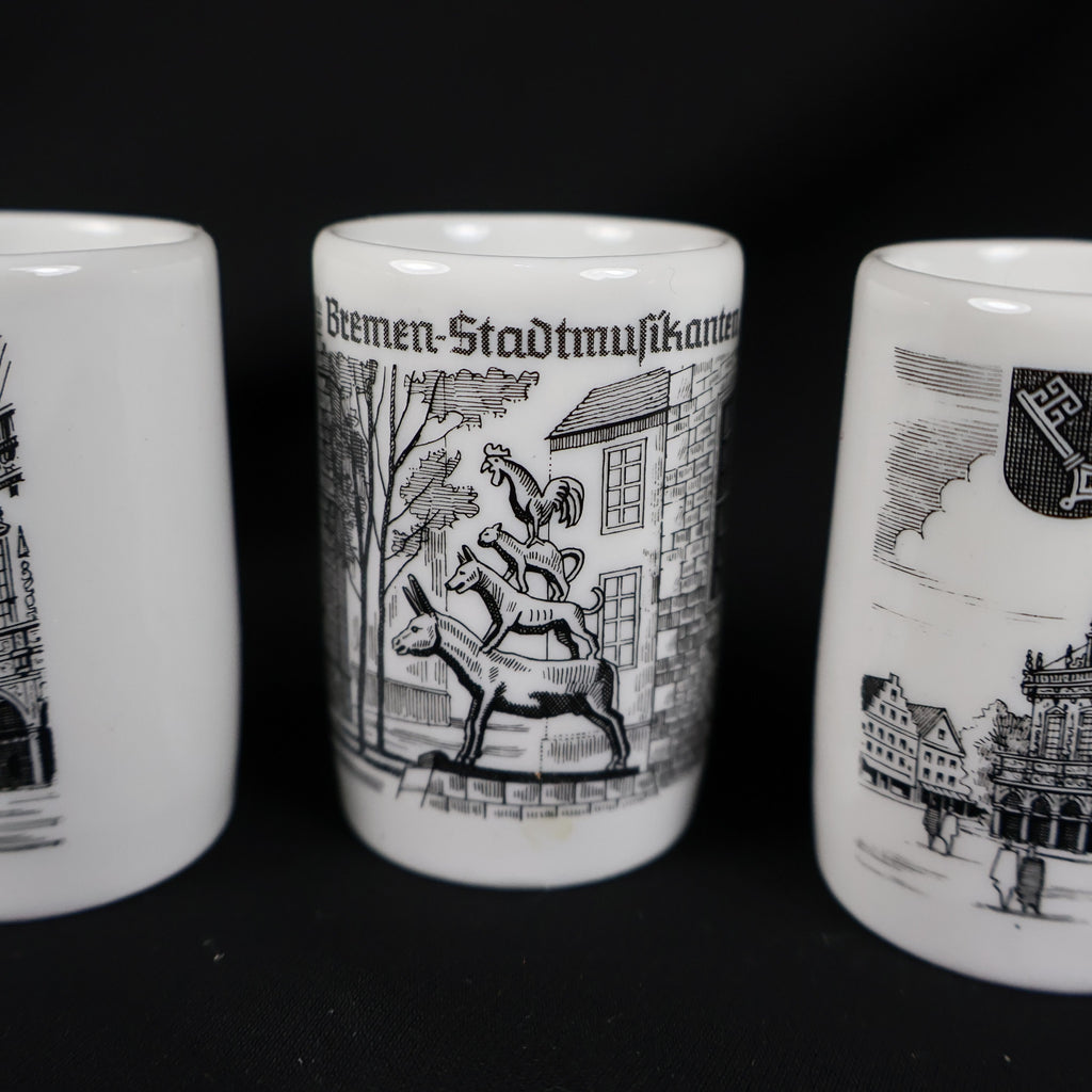 Detailed close-up of the Bremen Stadtmusikanten souvenir cup positioned between portions of the other two cups. The black illustration depicts a donkey, dog, cat, and rooster stacked together before buildings, with the German landmark title printed along the top.