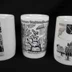 Detailed close-up of the Bremen Stadtmusikanten souvenir cup positioned between portions of the other two cups. The black illustration depicts a donkey, dog, cat, and rooster stacked together before buildings, with the German landmark title printed along the top.